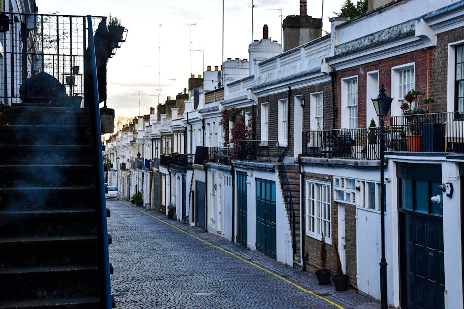 A row of terraced houses on a narrow cobbled street in a residential area during daylight, with several front doors, some with small potted plants, and black metal railings along the walkways. The street is used for home relocations and furniture transport, with a black van partially visible in the background. On the left side, a metal staircase with closed steps and a safety railing leads from an upper level to the street, which is lined with curb markings. The scene captures the typical setting for house removals in a historic district, where moving services involve loading furniture, boxes, and appliances through front doors or staircases, with some items possibly being wrapped in blankets or plastic for protection. The sky is partly cloudy, and the lighting highlights the details of the brickwork and architectural features of the buildings, reflecting the logistics involved in packing and moving within a densely built urban environment. Movers Highbury operates in such areas, offering professional removals services, including packing, loading, and transportation.