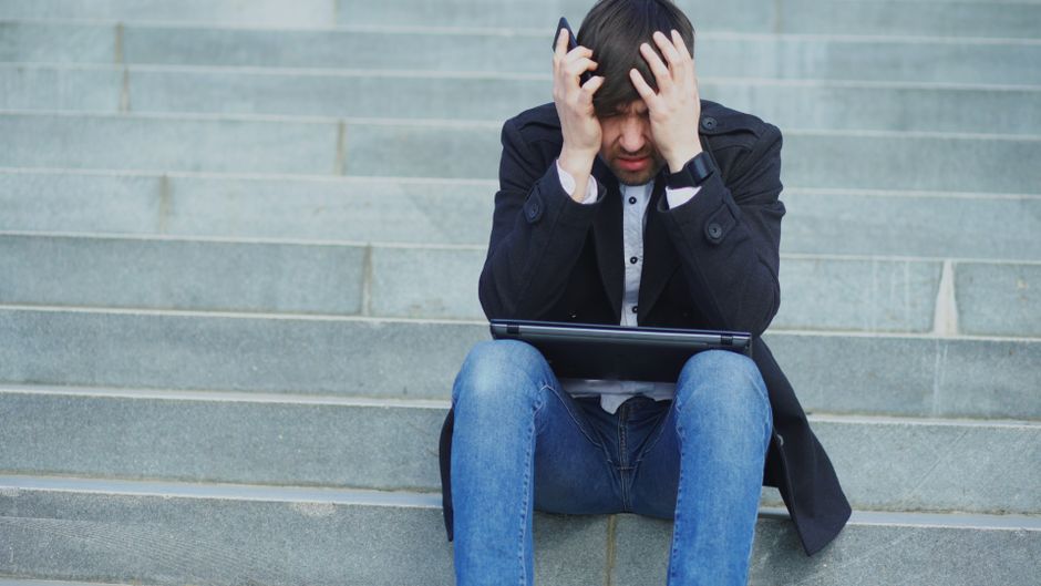 A young man with dark hair, wearing a black jacket, light blue shirt, and blue jeans, sitting on grey concrete steps outdoors. He is holding his head with both hands, appearing distressed and confused, with a laptop resting on his lap. The background shows the steps extending upwards with soft natural lighting. This image relates to common problems during house removals or home relocation, illustrating potential stress and frustration experienced during packing or moving logistics, as depicted in the context of Movers Highbury's services.