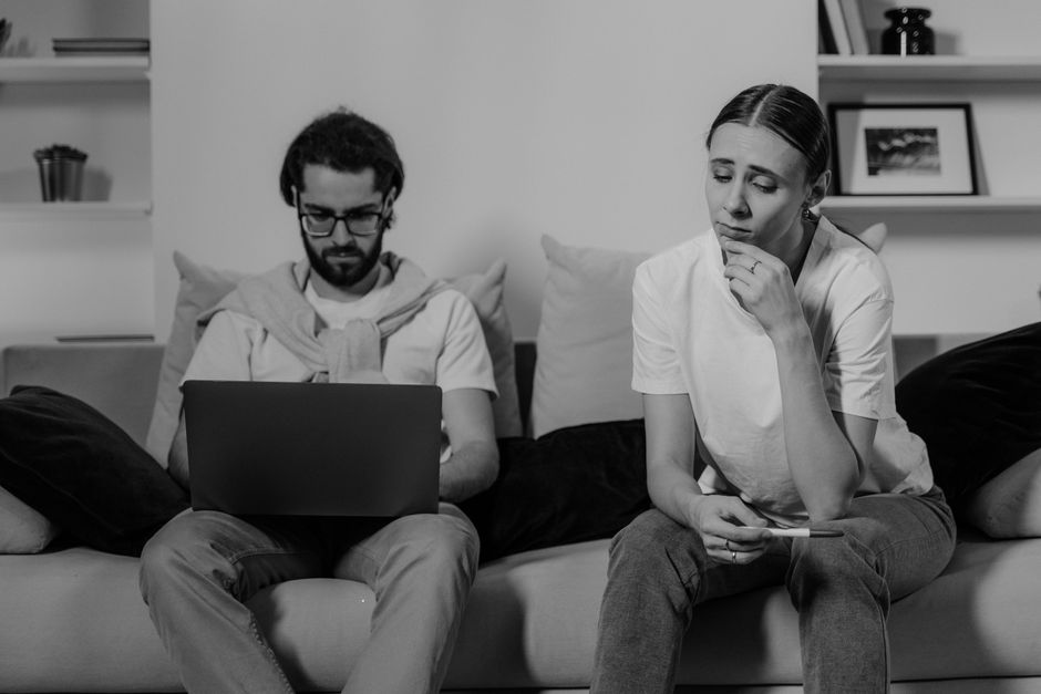 A black and white photograph showing a man and a woman seated on a sofa in a modern living room, involved in a stressful situation related to house removals or moving logistics. The man, with glasses and a beard, is focused on a laptop resting on his lap, with a sweater draped over his shoulders. He is wearing a light-colored shirt and jeans, sitting next to the woman. The woman has her hair pulled back and is dressed in a light t-shirt and jeans; she appears anxious or concerned, with her chin resting on her hand and a mobile phone in her other hand. Behind them, a wall-mounted shelf displays a framed photograph, a decorative object, and a couple of books, indicating a typical home environment. The sofa is decorated with cushions, and there are small items on a nearby table. The scene captures the tension and complexity often associated with home relocation, packing, and moving planning, as it reflects the stress experienced during the moving process, which the professional team at Movers Highbury helps to alleviate.