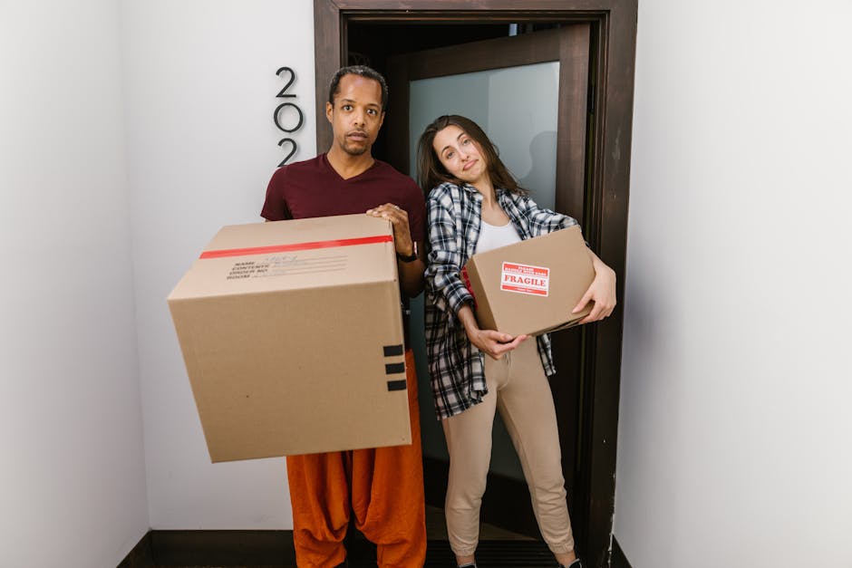 Two movers from Movers Highbury stand inside a narrow doorway of a residential building, each holding a cardboard box related to home relocation. The male mover, positioned on the left, is wearing a maroon t-shirt and orange trousers, and is carrying a large cardboard box sealed with red tape. The female mover, on the right, is dressed in a checkered shirt, white top, and beige trousers, and holds a smaller box labeled 'Fragile.' They are standing on a black floor with a plain white wall behind them, and a dark brown door frame with the building's street number partially visible. The scene illustrates the logistics of furniture transport and packing during a house move, involving careful handling of items within a confined entrance space, which may require specialized loading techniques. Movers Highbury is involved in coordinating the loading process, often requiring equipment such as trolleys or straps to ensure safe transport of belongings through difficult access points in Highbury estates.