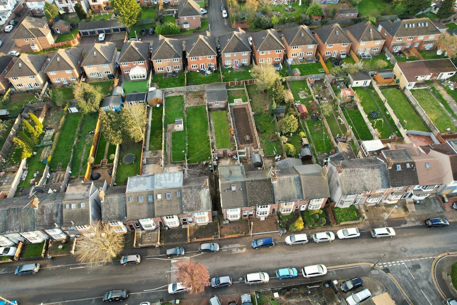 An aerial view of a row of terraced houses in a residential neighbourhood, with each property featuring small front gardens, driveways, and backyards. The back gardens are visible with various features such as lawn areas, garden sheds, patios, and planting beds, some enclosed with wooden or brick fences. In the foreground, a street lined with parked cars runs parallel to the houses. The image shows a moving process involving the loading of furniture and boxes into a large van parked on the street, with movers using trolleys and protective blankets to transport household items. Cardboard boxes, wrapped furniture, and packing materials are visible, indicating a home relocation. The environment is well-lit with natural daylight, and the surrounding greenery includes trees and shrubs, adding to the residential setting. This scene exemplifies the logistical aspects of house removals on estates where access may be challenging, highlighting the procedures managed by [COMPANY_NAME], such as furniture transport and packing during the difficult access move in Highbury.