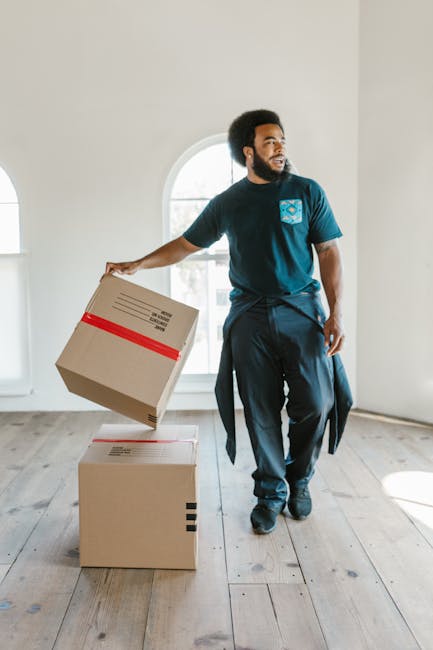 A person dressed in a grey sweater is in the process of sealing a cardboard moving box with packing tape, which bears the logo of Movers Highbury. The individual is holding the tape dispenser in their
