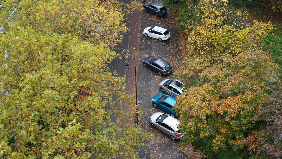 An aerial view in black and white showing a large parking lot filled with numerous parked vehicles, with some leaving spaces for new arrivals. The parking area is adjacent to a residential or commerci