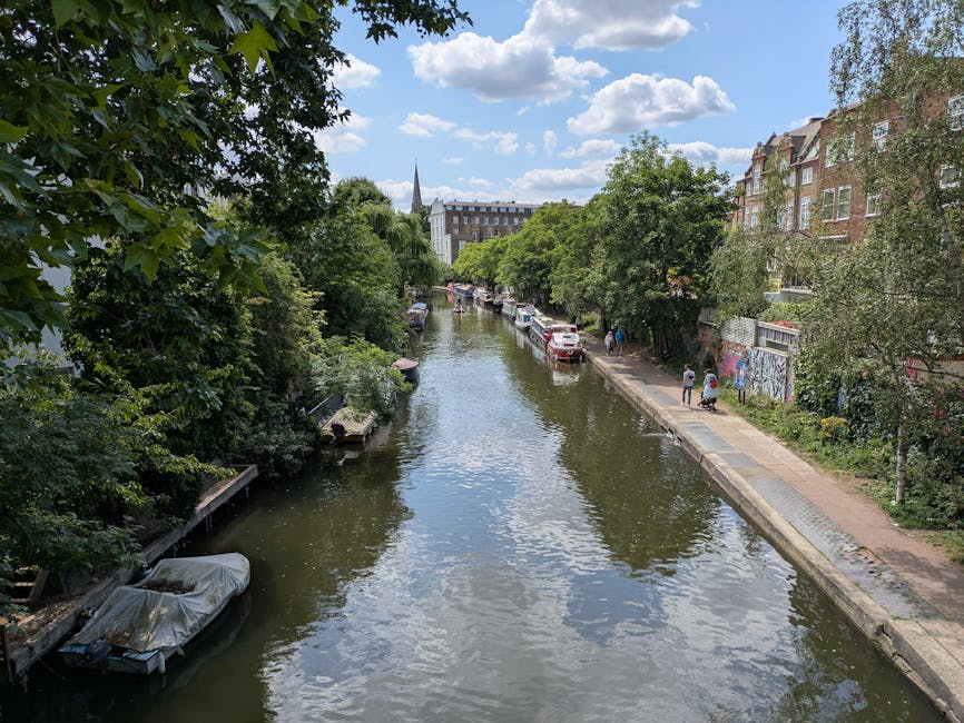 A narrow canal running through a residential area on a bright, partly cloudy day. On the left side, lush green trees overhang the water, with a small boat docked along the canal edge, partially covered with a protective tarp. On the right side, a paved walking path runs parallel to the canal, with a few pedestrians visible and parked cars lining the street further from the water. Residential buildings with brick facades and large windows are situated behind the trees and along the sidewalk. A church steeple rises in the background above the cityscape. The scene captures the tranquil environment typical of a home relocation or moving logistics setting in Highbury, with the canal and surroundings emphasizing the local neighbourhood atmosphere.