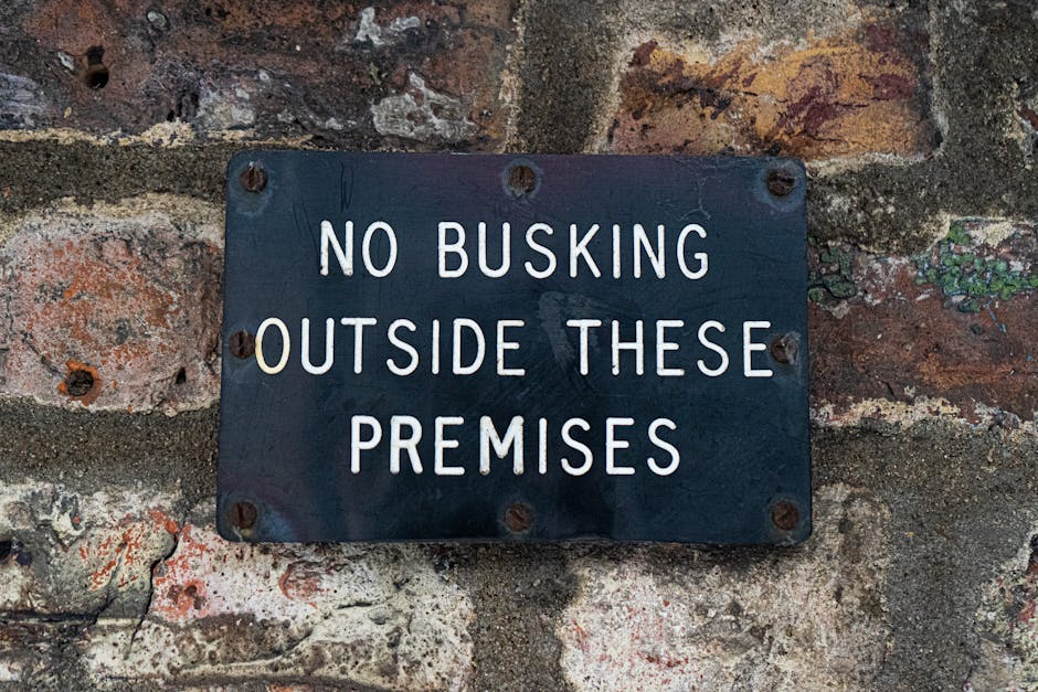 A rectangular metal sign with a dark background and white uppercase lettering mounted on an aged brick wall, displaying the message 'NO BUSKING OUTSIDE THESE PREMISES.' The sign is secured with four rusted screws, one at each corner. The brick wall behind shows a weathered texture with various shades of red, brown, and some patches of moss or lichen growth. The sign is positioned at about eye level, and its purpose appears to be to prohibit street performances or busking in the specific area. This setting is likely near a building entrance or a part of a property where quiet or restricted activity is required, relevant to house removals or relocation contexts that involve understanding local regulations for moving in or out of premises. The overall scene conveys a sense of compliance and respect for property boundaries during planning logistics, such as relocation or furniture transport, which is relevant to Movers Highbury's services on the page about Islington Council permit rules for Highbury removals.
