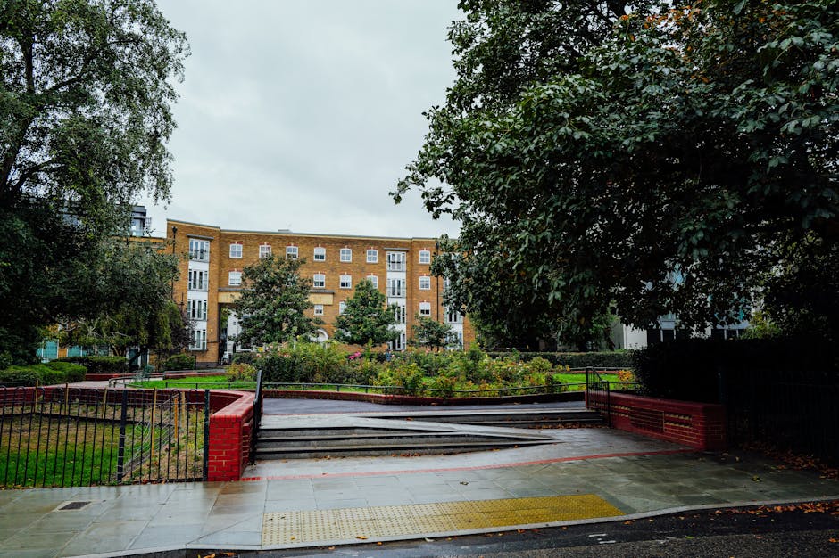 A view of a residential apartment building with multiple stories and white-framed windows, situated behind a small park with green grass, trees, and flower beds. In the foreground, a concrete pathway featuring a tactile paving surface for accessibility leads to a flight of black metal stairs with red brick end posts, which ascend into a building's entrance. On the left side, a black metal fence borders the pathway, while on the right, a large leafy tree partially shades the area. The scene is overcast with grey skies, and the environment suggests a peaceful urban residential area. The image is associated with house removals and relocation services, as provided by Movers Highbury, and illustrates the logistical considerations of navigating stairs during furniture transport or home moving processes.