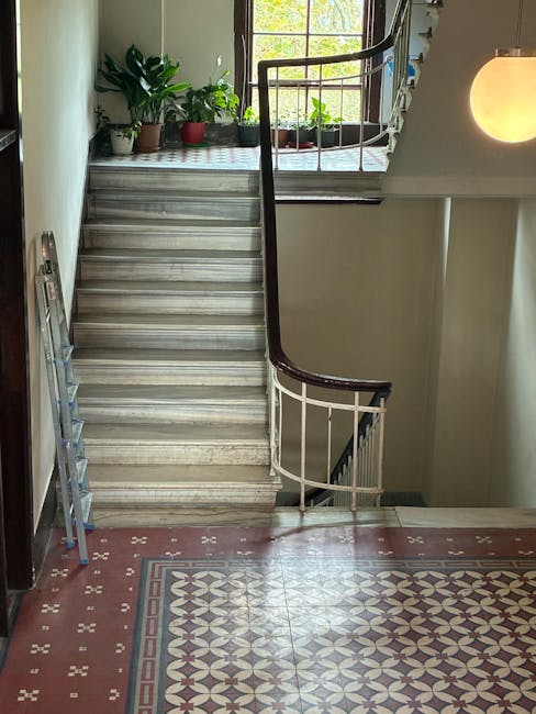 A photograph of an indoor staircase within a residential property, showing a wooden handrail and white metal balustrades. The staircase has marble steps leading from the ground floor, with some potted plants, including a large green leafy plant and smaller plants, placed on a window sill at the top of the stairs. Natural light filters through a window with multiple panes, illuminating the landing and highlighting the indoor plants. To the left of the stairs, a teal and silver folding ladder is leaning against the wall. The floor at the bottom of the staircase features patterned ceramic tiles with a geometric design in shades of red, white, and gray. A round wall-mounted light fixture emits a warm glow on the right side of the image. This scene illustrates a typical domestic staircase area, captured during the home relocation process by Movers Highbury, with visible elements related to furniture transport and packing preparation for house removals.