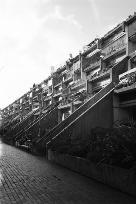 A view of a residential apartment building with multiple stories and white-framed windows, situated behind a small park with green grass, trees, and flower beds. In the foreground, a concrete pathway featuring a tactile paving surface for accessibility leads to a flight of black metal stairs with red brick end posts, which ascend into a building's entrance. On the left side, a black metal fence borders the pathway, while on the right, a large leafy tree partially shades the area. The scene is overcast with grey skies, and the environment suggests a peaceful urban residential area. The image is associated with house removals and relocation services, as provided by Movers Highbury, and illustrates the logistical considerations of navigating stairs during furniture transport or home moving processes.
