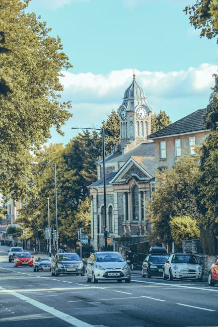 A row of parked cars on a street in front of a historic building with a clock tower, surrounded by lush green trees under a bright blue sky with some clouds. The scene captures the urban environment typical of a residential or mixed-use area in Highbury, with trees providing shade along the pavement. The image offers a clear view of moving and parking activity, which may be relevant to house removals and furniture transport services provided by Movers Highbury, especially during home relocation or packing and moving processes in Canonbury Road and Highbury. The streetlights and modern vehicle designs contrast with the older architectural style of the building, emphasizing the blend of historic and contemporary elements common in the area.