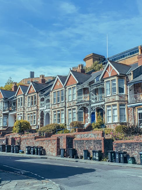 A row of historic Victorian-style terraced houses with red brick facades and decorative white trim, situated on a street with a clear blue sky overhead. The buildings feature large bay windows, some with ornate ironwork balconies, and small front gardens enclosed by low brick walls. The street has a paved sidewalk with black wheelie bins lined up along the curb, and the scene is illuminated by natural daylight, emphasizing the architectural details. As part of professional removals services, Movers Highbury may coordinate the careful packing and loading of furniture and boxes from such properties during home relocation or moving logistics, involving careful transportation of household items in a high-demand urban environment.