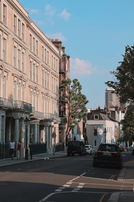 A row of parked cars on a street in front of a historic building with a clock tower, surrounded by lush green trees under a bright blue sky with some clouds. The scene captures the urban environment typical of a residential or mixed-use area in Highbury, with trees providing shade along the pavement. The image offers a clear view of moving and parking activity, which may be relevant to house removals and furniture transport services provided by Movers Highbury, especially during home relocation or packing and moving processes in Canonbury Road and Highbury. The streetlights and modern vehicle designs contrast with the older architectural style of the building, emphasizing the blend of historic and contemporary elements common in the area.