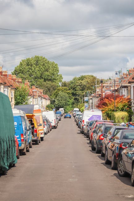 A residential street scene showing parked cars lining both sides of the road, with a variety of vehicles including vans and small cars, some with cardboard and plastic covers, indicating ongoing home relocation activities. The street is flanked by terraced houses with brick facades, small front gardens, and mature trees providing greenery. Overhead, electrical wires stretch across the sky, which is partly cloudy with patches of blue. Toward the far end of the street, a moving company van from Movers Highbury is visible, positioned near the pavement, suggesting that furniture transport and packing and moving processes are underway as part of a house removal service near Highbury Fields for narrow access homes.