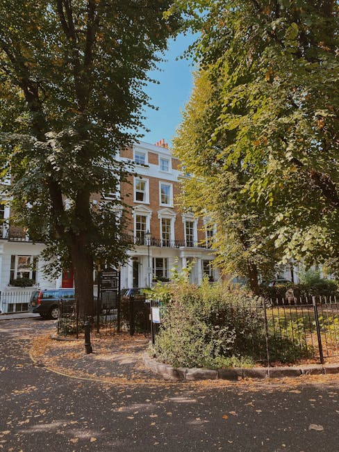 A multi-storey residential building with a white facade and large windows, partially obscured by tall, leafy trees with green and yellow foliage, indicating an autumn setting. The building is situated on a corner with a small front garden enclosed by a black metal fence, and a paved pathway leading to the entrance. In front of the building, on the pavement, there is a parking area with a grey vehicle, and the surrounding area is shaded by the trees, creating a contrast between sunlight and shadow. The scene is part of a typical urban street environment, with the appearance of a house removal or moving process supported by the nearby greenery and accessible street. This setting is consistent with the context of house removals near Highbury Fields, as offered by Movers Highbury, highlighting the typical environment for relocating furniture and boxes through narrow access homes.