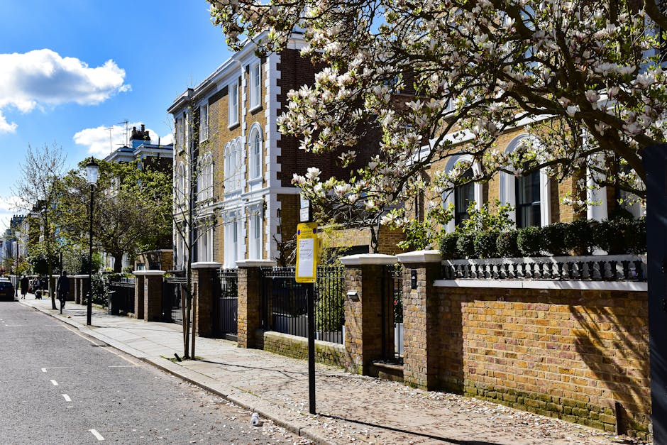 A residential street scene showing parked cars lining both sides of the road, with a variety of vehicles including vans and small cars, some with cardboard and plastic covers, indicating ongoing home relocation activities. The street is flanked by terraced houses with brick facades, small front gardens, and mature trees providing greenery. Overhead, electrical wires stretch across the sky, which is partly cloudy with patches of blue. Toward the far end of the street, a moving company van from Movers Highbury is visible, positioned near the pavement, suggesting that furniture transport and packing and moving processes are underway as part of a house removal service near Highbury Fields for narrow access homes.